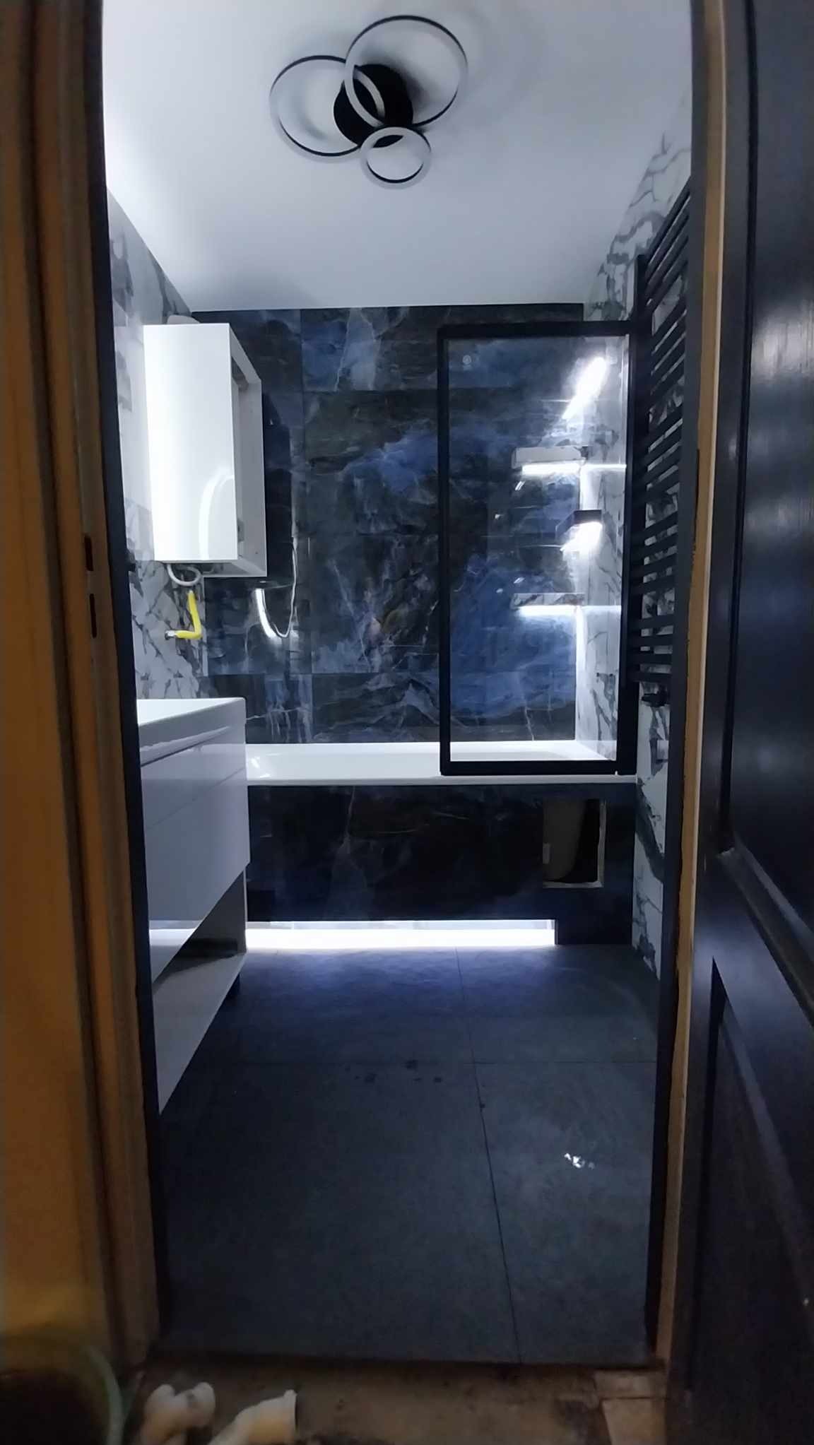 Nowoczesna łazienka z podświetleniem LED pod wanną, marmurowymi płytkami w odcieniach granatu i bieli oraz czarną ramą kabiny prysznicowej i grzejnikiem drabinkowym, widok zza otwartych drzwi.