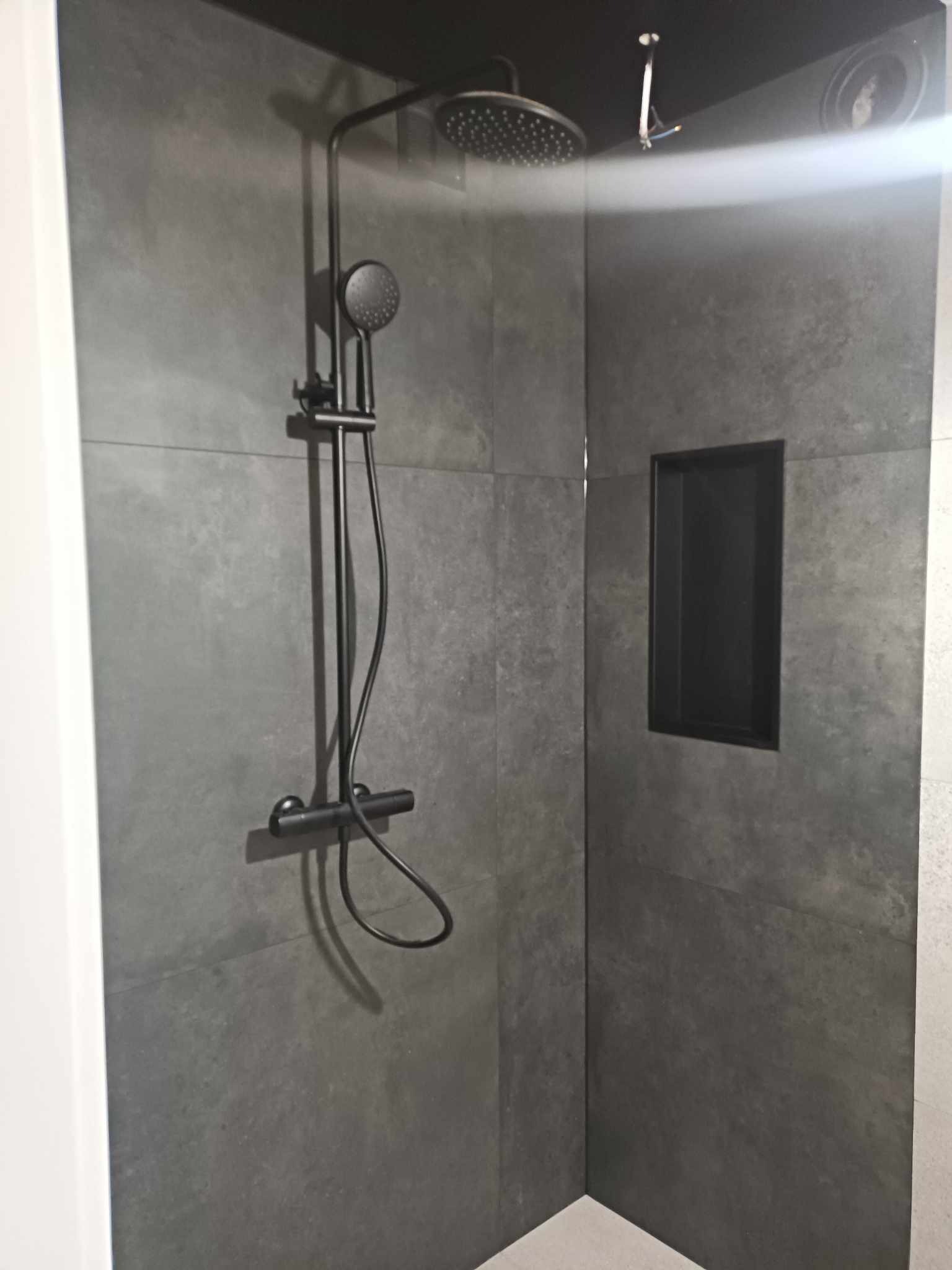 Nowoczesna kabina prysznicowa z czarną armaturą i wnęką w ścianie wyłożonej szarymi płytkami imitującymi beton.