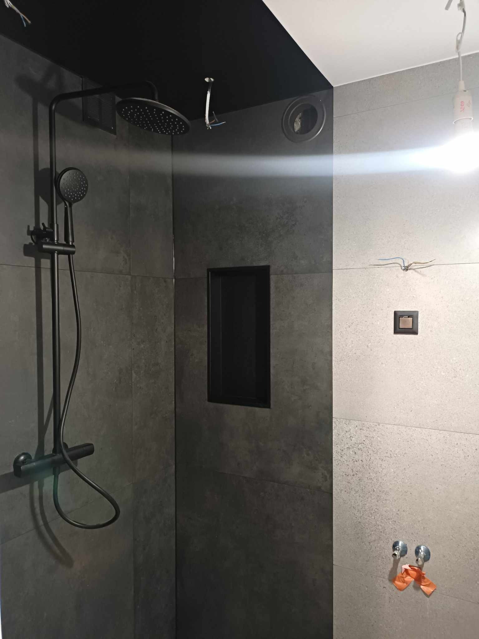 Częściowo wykończona łazienka z czarną armaturą, wnęką w ścianie i widocznymi przewodami elektrycznymi.