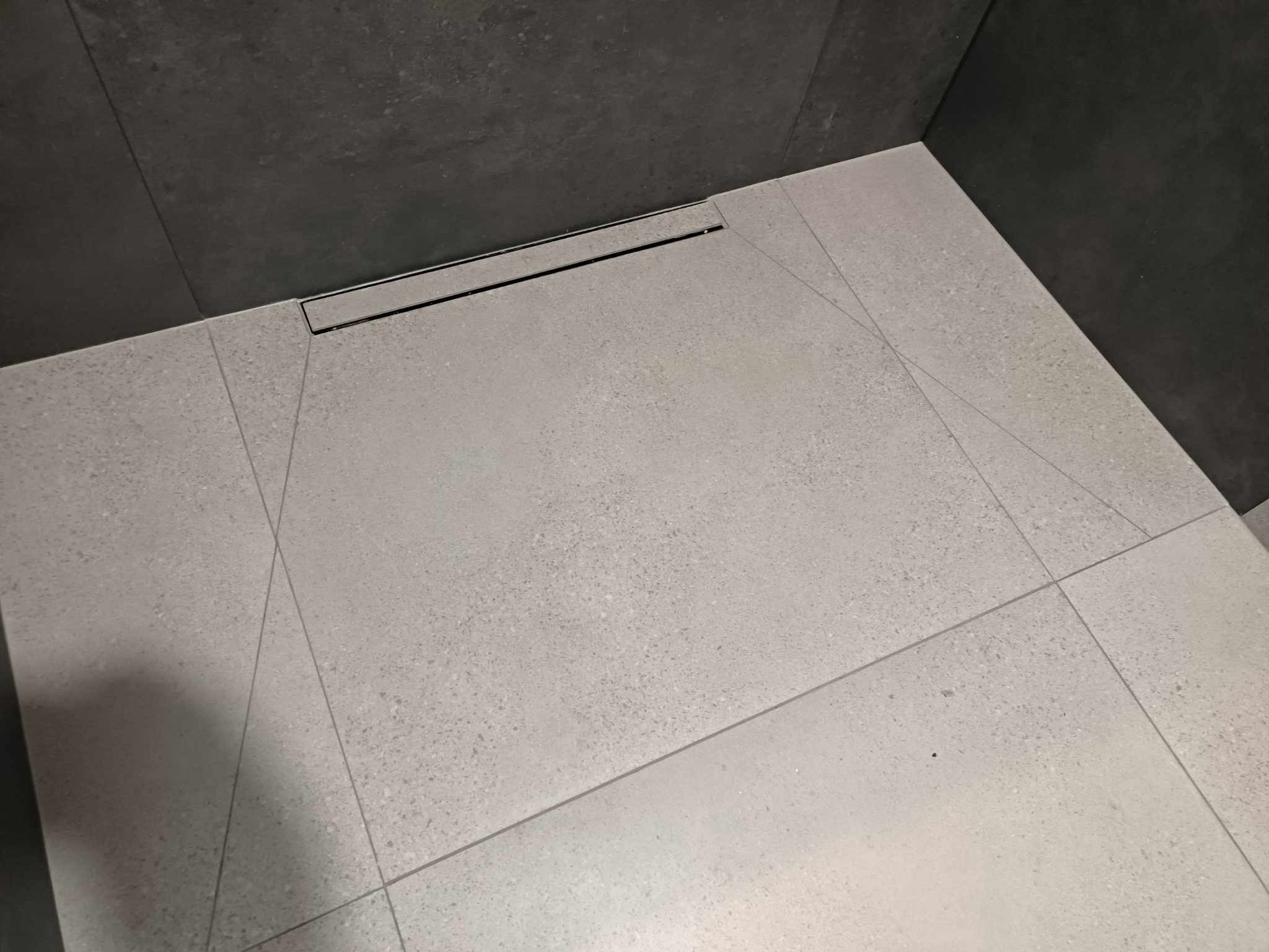 Nowoczesna kabina prysznicowa walk-in z odpływem liniowym i szarymi płytkami na ścianach i podłodze, ułożonymi w geometryczny wzór.