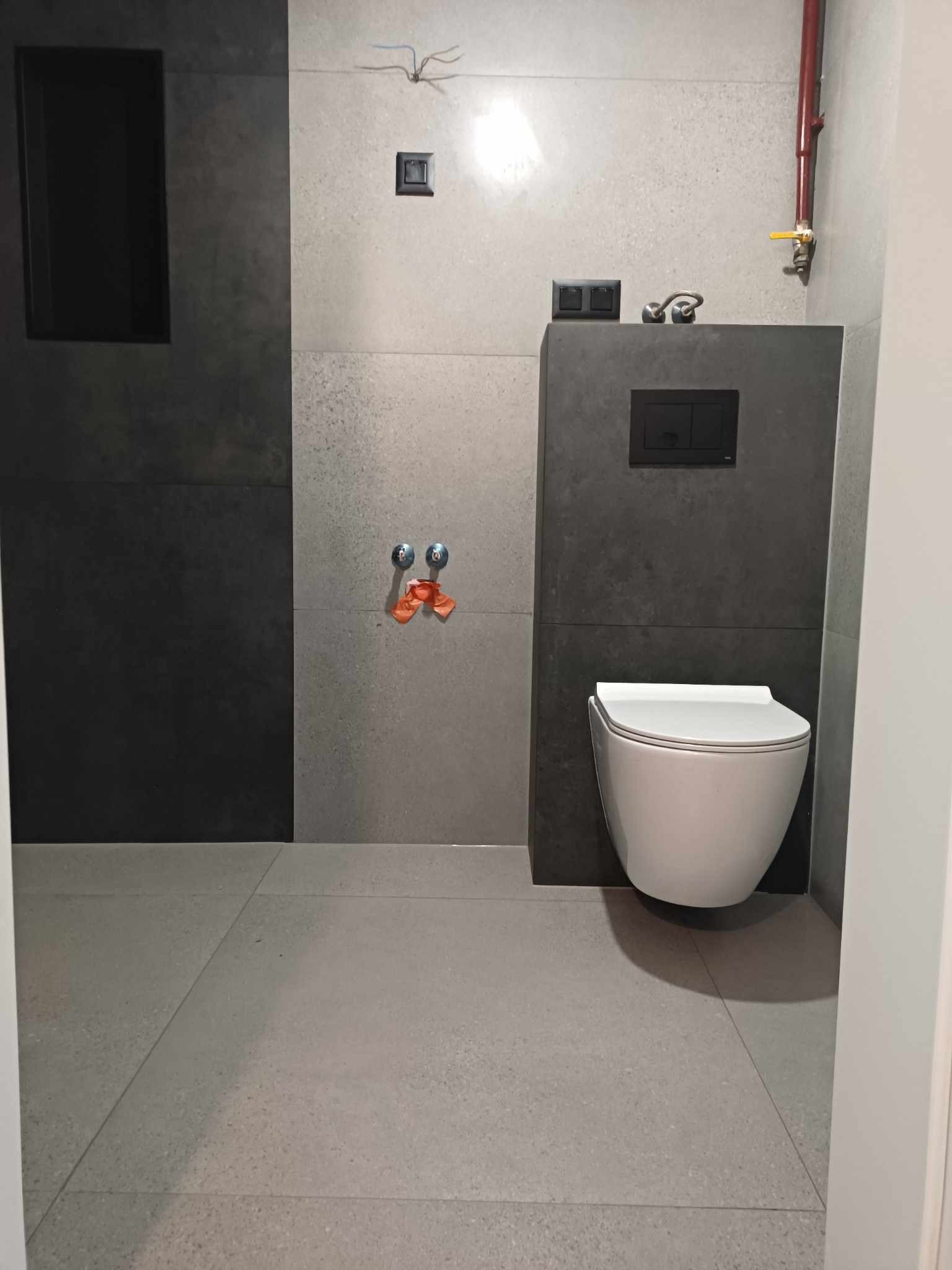 Nowoczesna łazienka w szarościach, z wiszącą toaletą, widoczne przyłącza wodne i instalacja elektryczna, surowy styl.