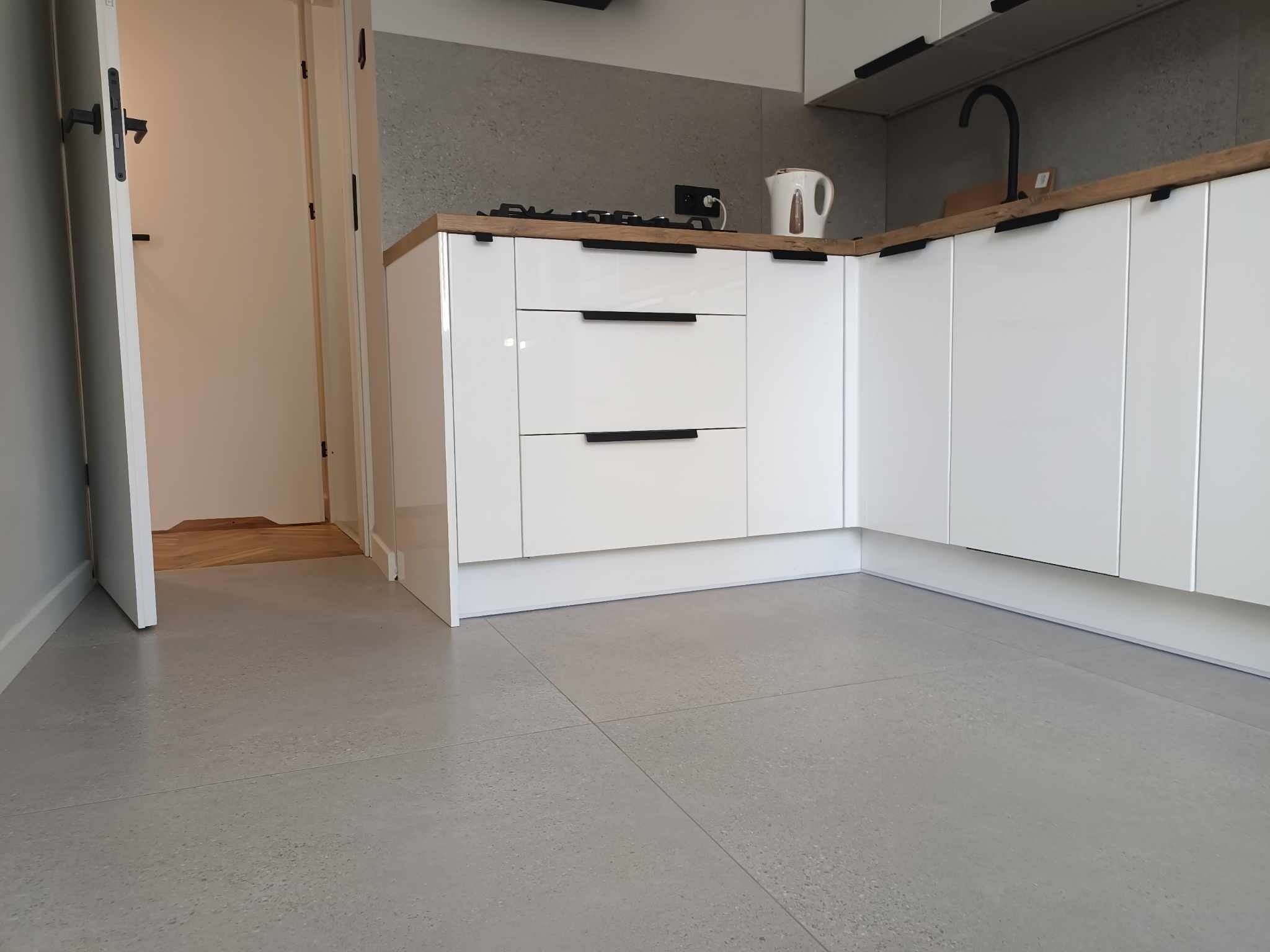 Nowoczesna kuchnia z białymi lakierowanymi szafkami, drewnianym blatem, czarną armaturą i szarą posadzką z płytek imitujących beton.