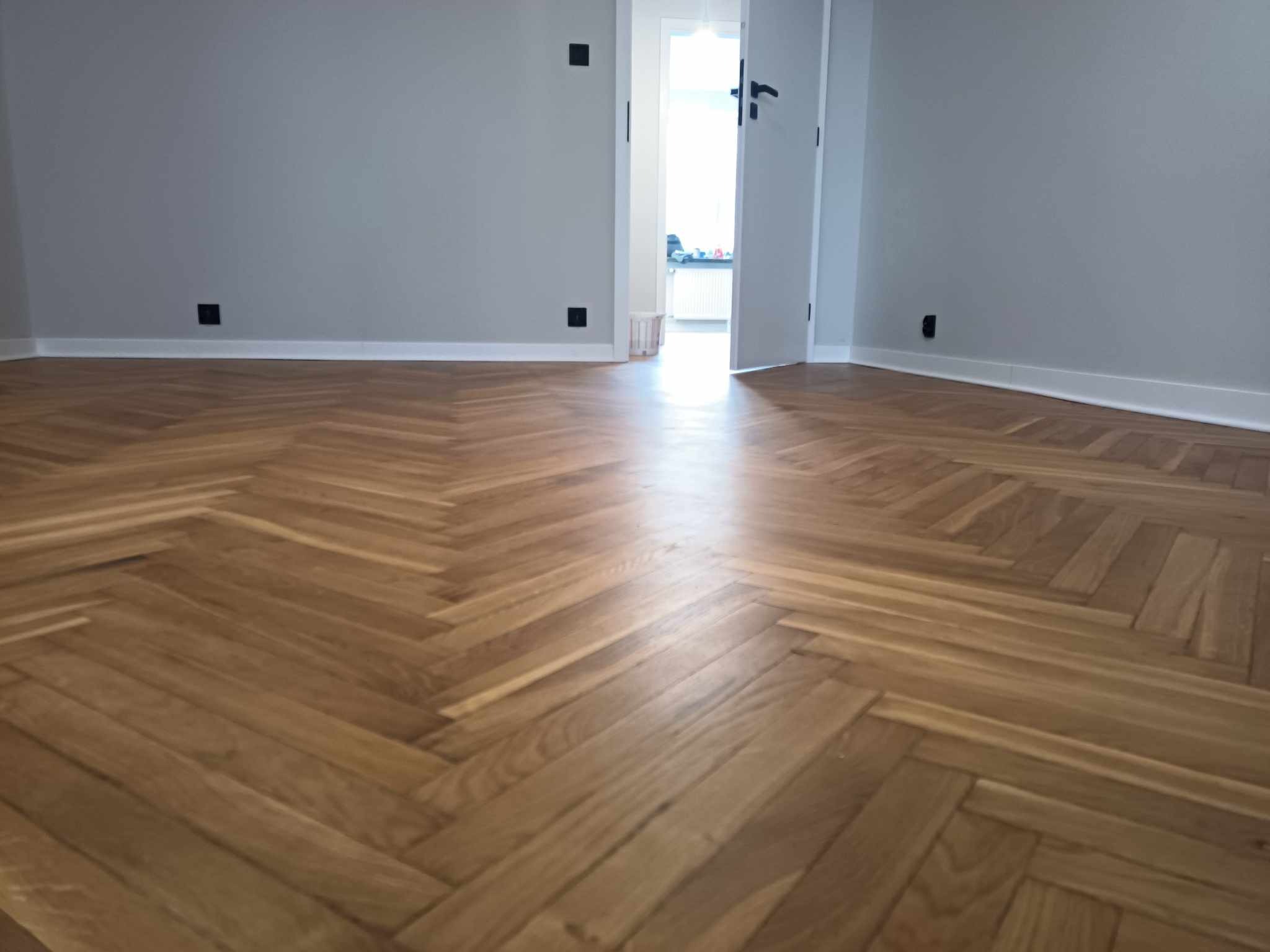 Ułożona parkietowa podłoga w jodełkę, szare ściany z czarnymi kontaktami, otwarte drzwi do jasnego pomieszczenia.