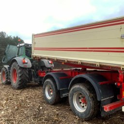 USŁUGI KOPARKO - ŁADOWARKĄ Jakub Kobuszewski - Ciężki traktor z otwartą maską i przyczepą na polu z resztkami po zbiorach, widoczne duże koła i błotniki przyczepy.