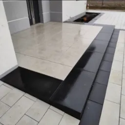 Nowoczesne wejście do domu z jasnej kostki brukowej z ciemnymi obrzeżami i schodami, widoczne odbicie światła na mokrej powierzchni.