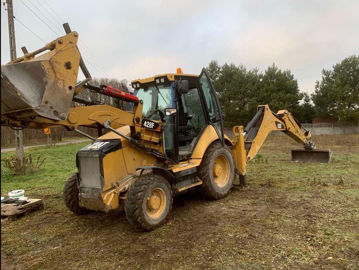 Żółta koparko-ładowarka Caterpillar 428F z otwartymi drzwiami kabiny, stojąca na grząskim terenie, z widocznym stosem palet w tle i rozłożonymi ramionami kopiącymi.