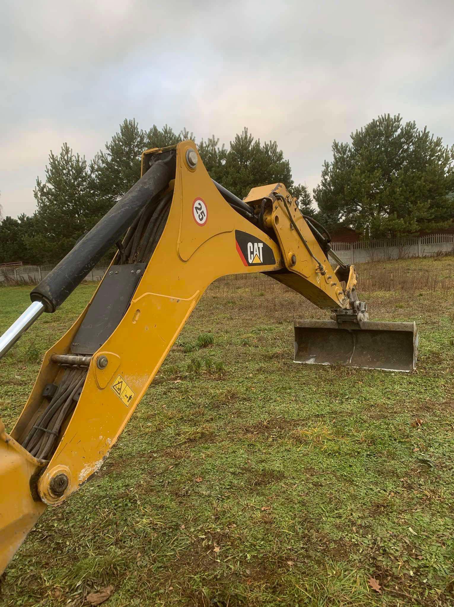 Żółty wysięgnik koparki CAT z łyżką na tle zielonej trawy i drzew, widok z boku.