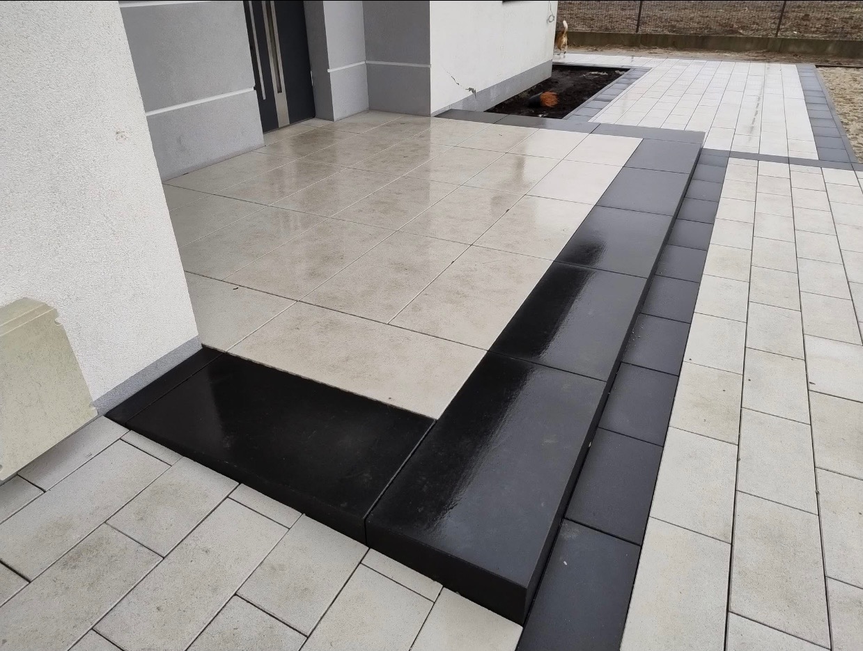 Nowoczesne wejście do domu z jasnej kostki brukowej z ciemnymi obrzeżami i schodami, widoczne odbicie światła na mokrej powierzchni.