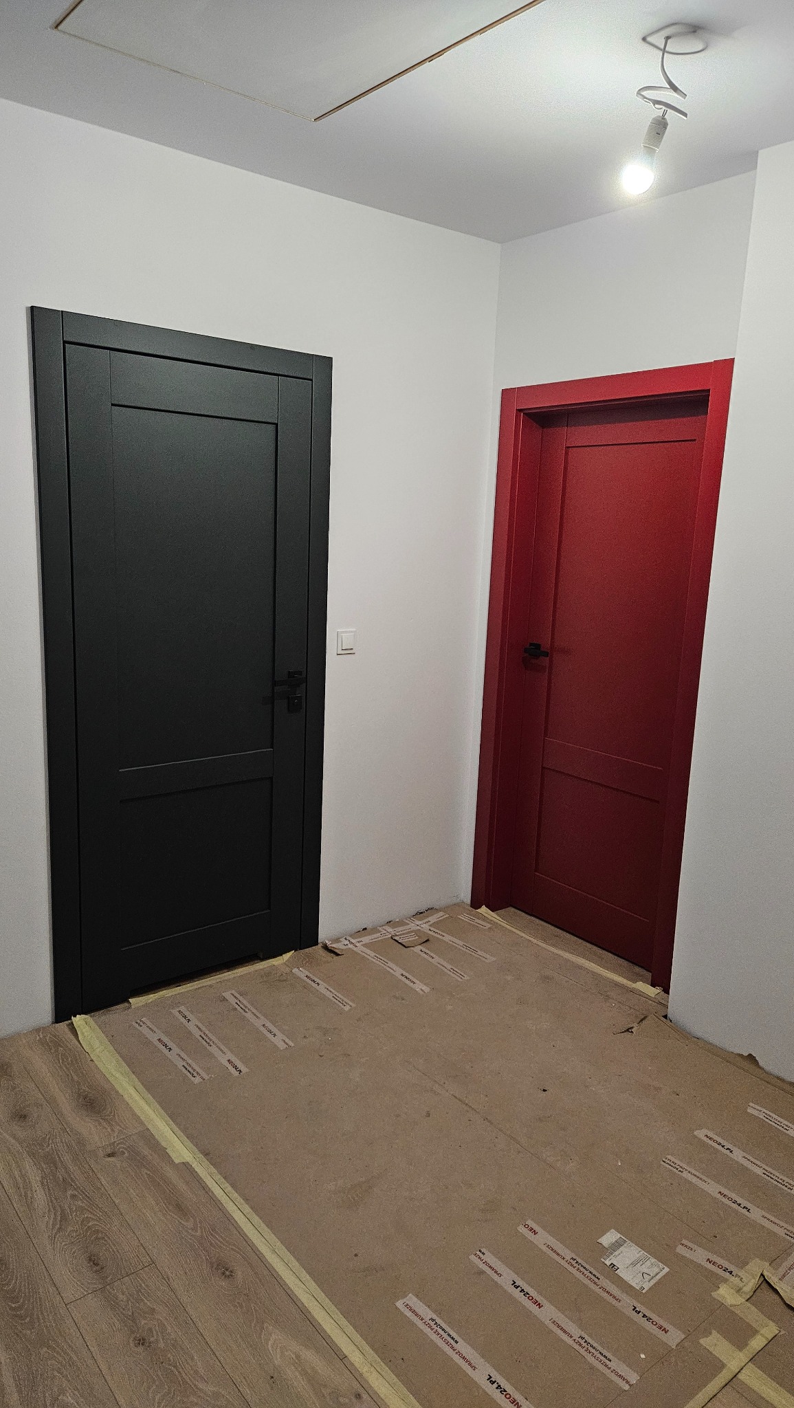 Dwa nowe, pomalowane skrzydła drzwiowe w kolorach czarnym i czerwonym, zamontowane w białym wnętrzu; podłoga zabezpieczona kartonem z widocznymi taśmami i napisami reklamowymi.