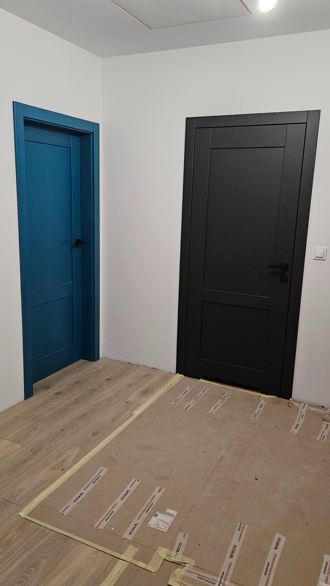 Dwa zamontowane, nowoczesne drzwi wewnętrzne: turkusowe i czarne, z widocznym zabezpieczeniem podłogi kartonem i taśmą malarską.