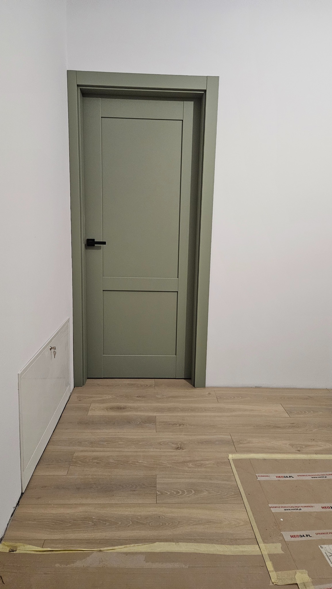 Zamknięte, dwupanelowe drzwi w odcieniu oliwkowej zieleni z czarną klamką, zamontowane w białej ścianie, z widoczną podłogą z paneli i zabezpieczeniem podłogi kartonem z nadrukiem reklamowym.