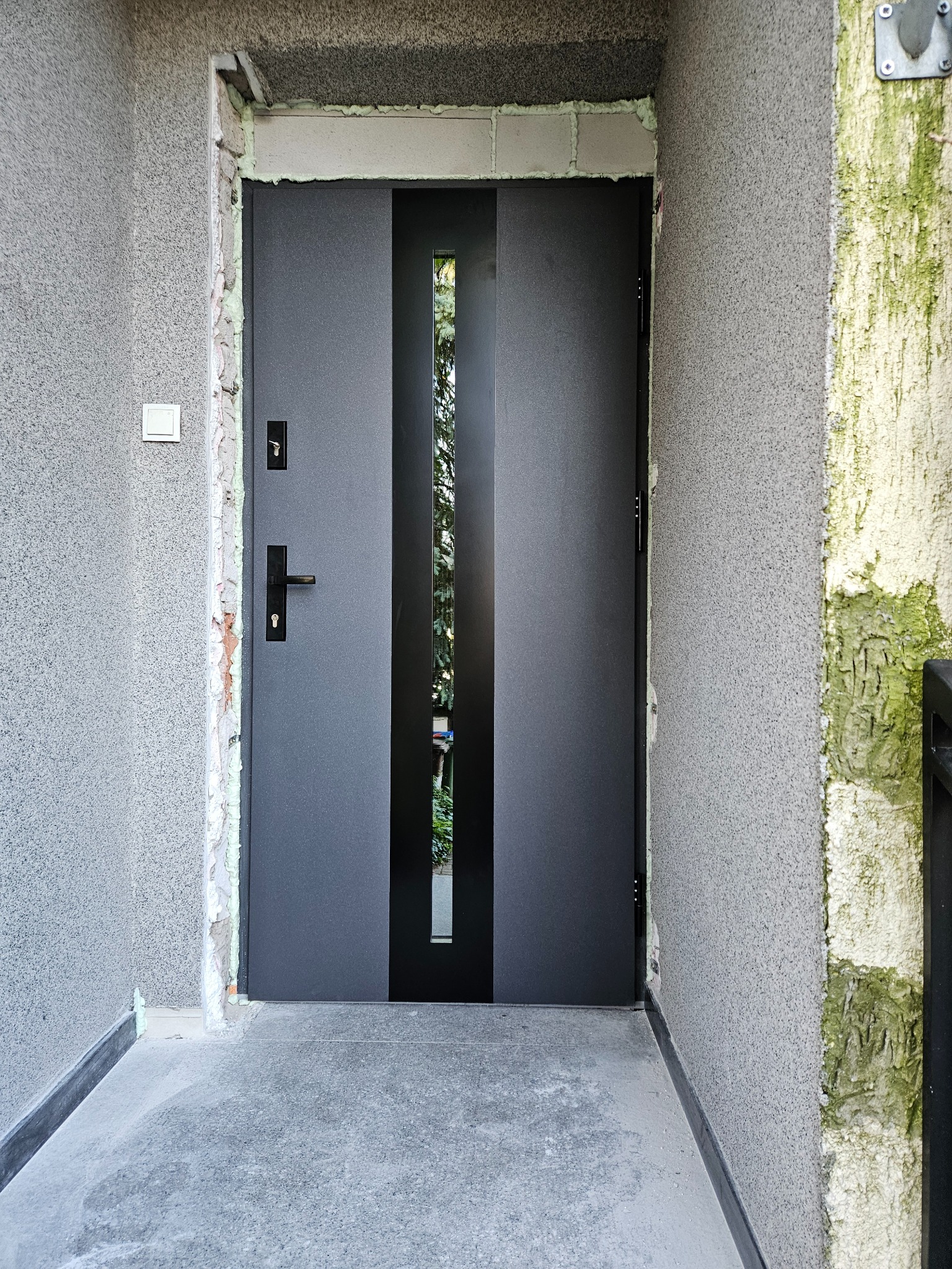 Szare drzwi wejściowe z czarnym pionowym przeszkleniem, zamontowane w surowym otworze, widoczna pianka montażowa i ślady po pracach wykończeniowych na szarej elewacji.