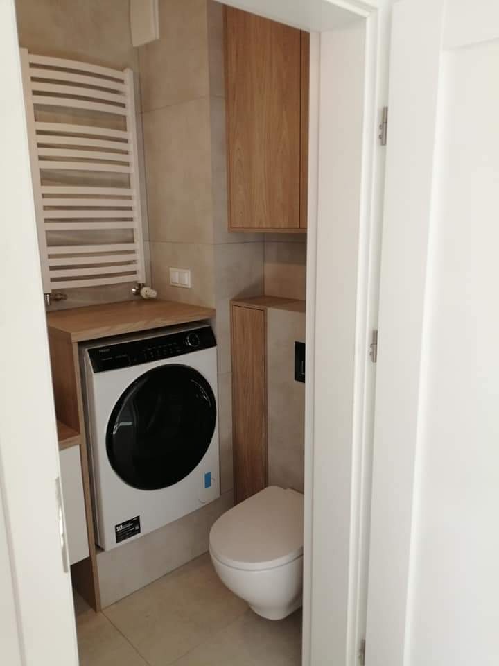 Mała łazienka z pralką zabudowaną pod drewnianym blatem, wiszącą szafką i grzejnikiem drabinkowym, widok zza uchylonych białych drzwi.