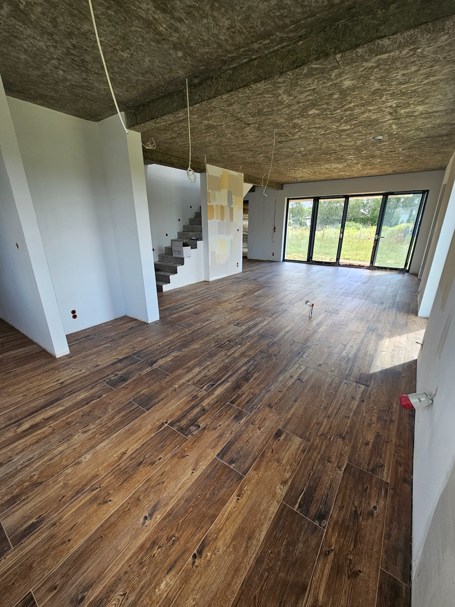 Wnętrze w trakcie wykańczania: brązowe płytki podłogowe imitujące drewno, surowy betonowy sufit, białe ściany z widocznymi przewodami elektrycznymi i fragmentem schodów z pomalowanymi próbkami...