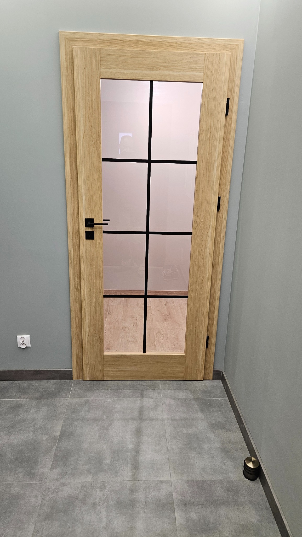 Drewniane drzwi wewnętrzne z szybami podzielonymi czarnymi szprosami, widoczne na tle szarej ściany i szarej podłogi z płytek, z czarną klamką i zawiasami.