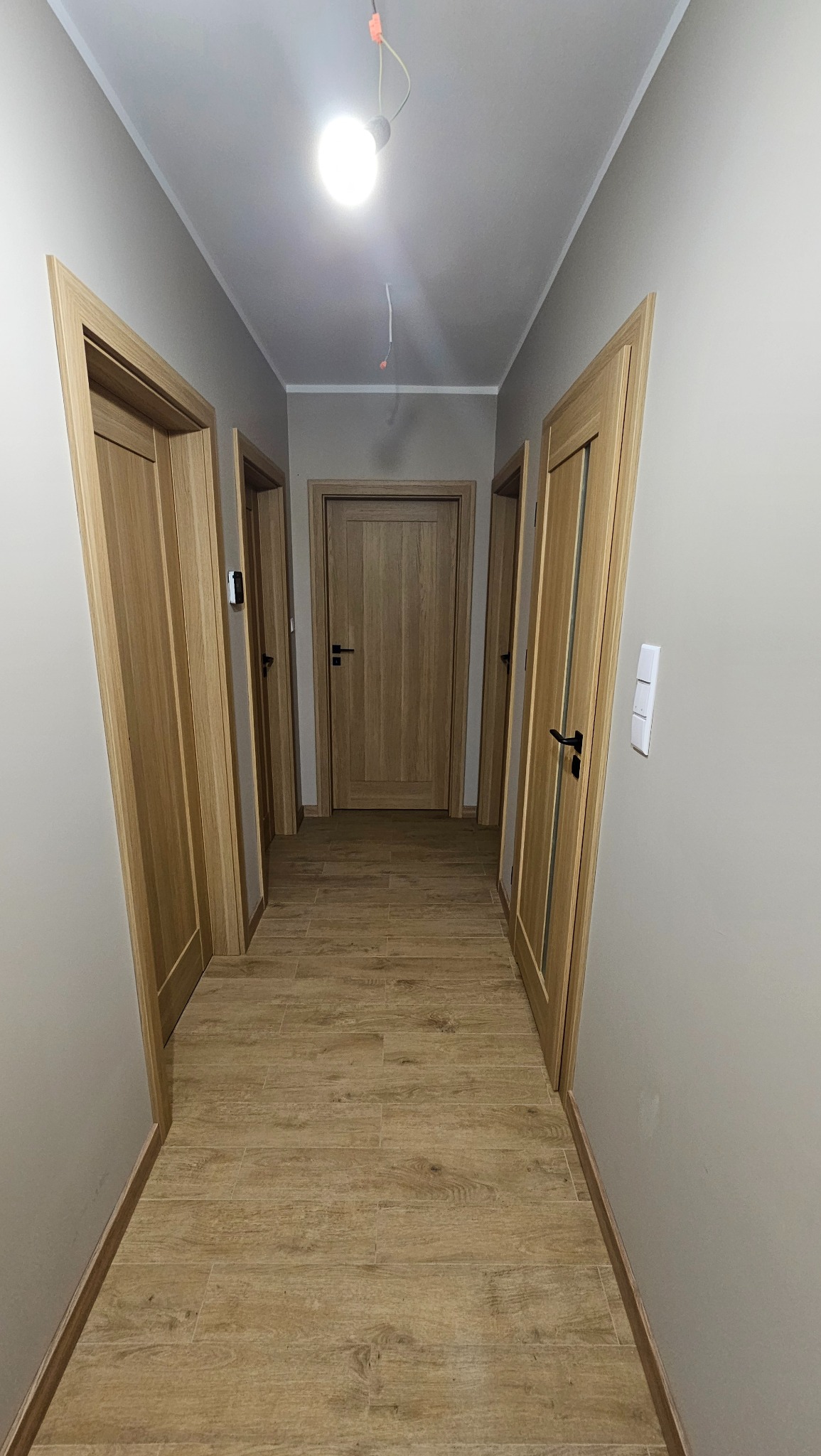 Korytarz z trzema drewnianymi drzwiami z czarnymi klamkami, wykończony płytkami imitującymi drewno, oświetlony pojedynczą żarówką.