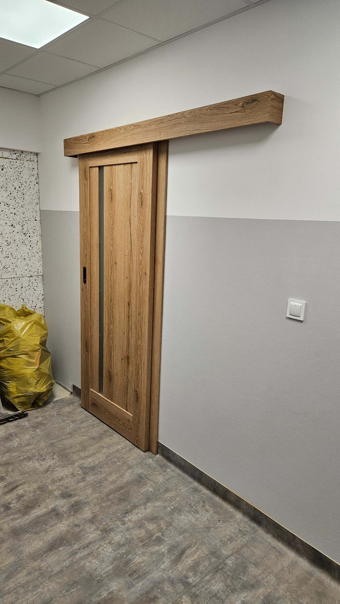 Zamontowane drewniane drzwi przesuwne z pionowym przeszkleniem, widoczne na tle dwukolorowej ściany i worka z odpadami budowlanymi.