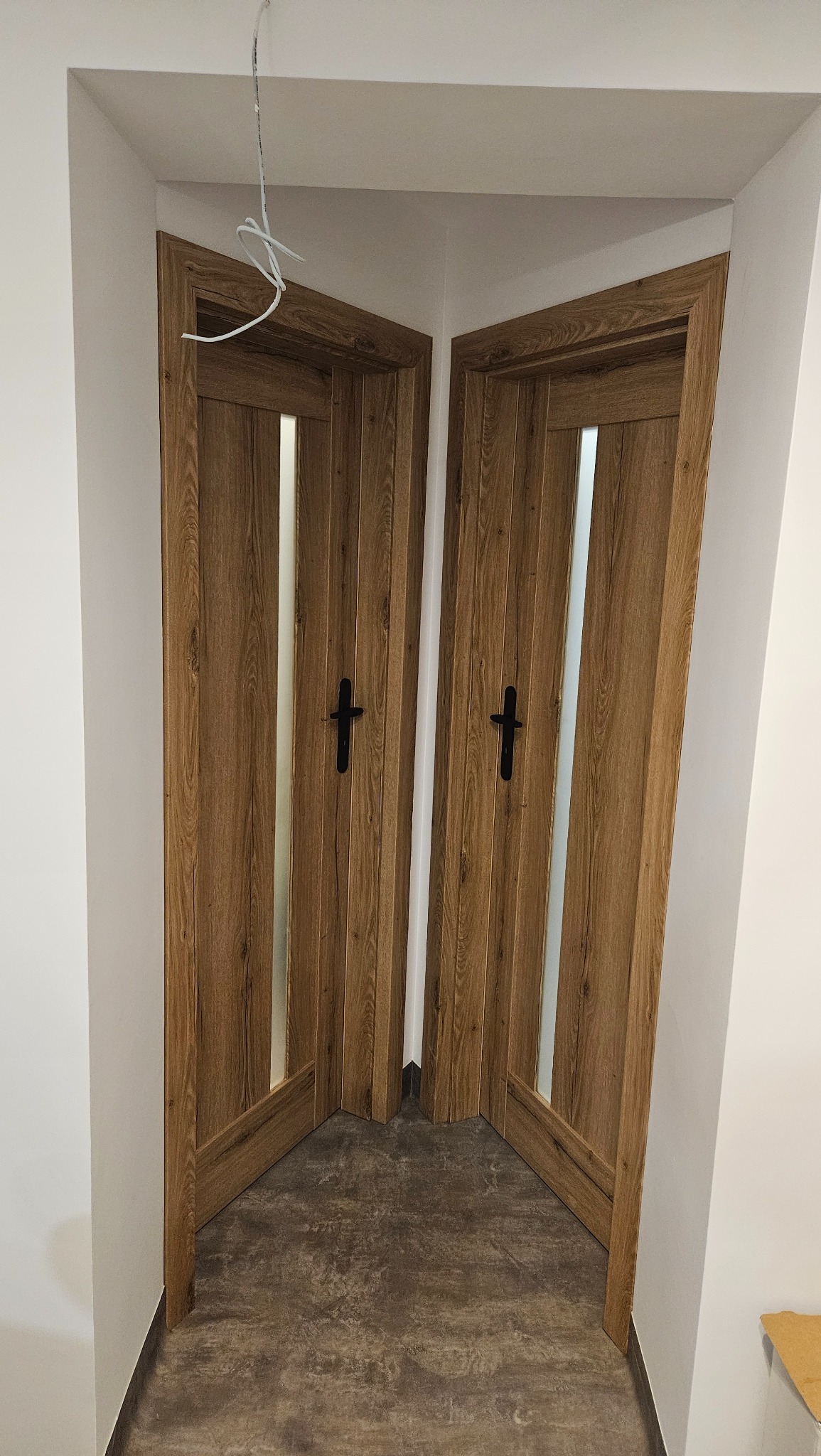 Dwa skrzydła drzwiowe z drewnianą okleiną i przeszkleniem, zamontowane w narożniku, widoczne przewody instalacji elektrycznej.