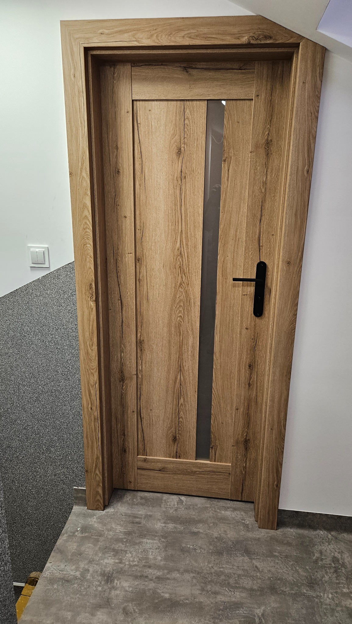 Drewniane drzwi wewnętrzne z czarną klamką i wąskim przeszkleniem, zamontowane w pomieszczeniu z szarą, teksturowaną ścianą i betonową podłogą.