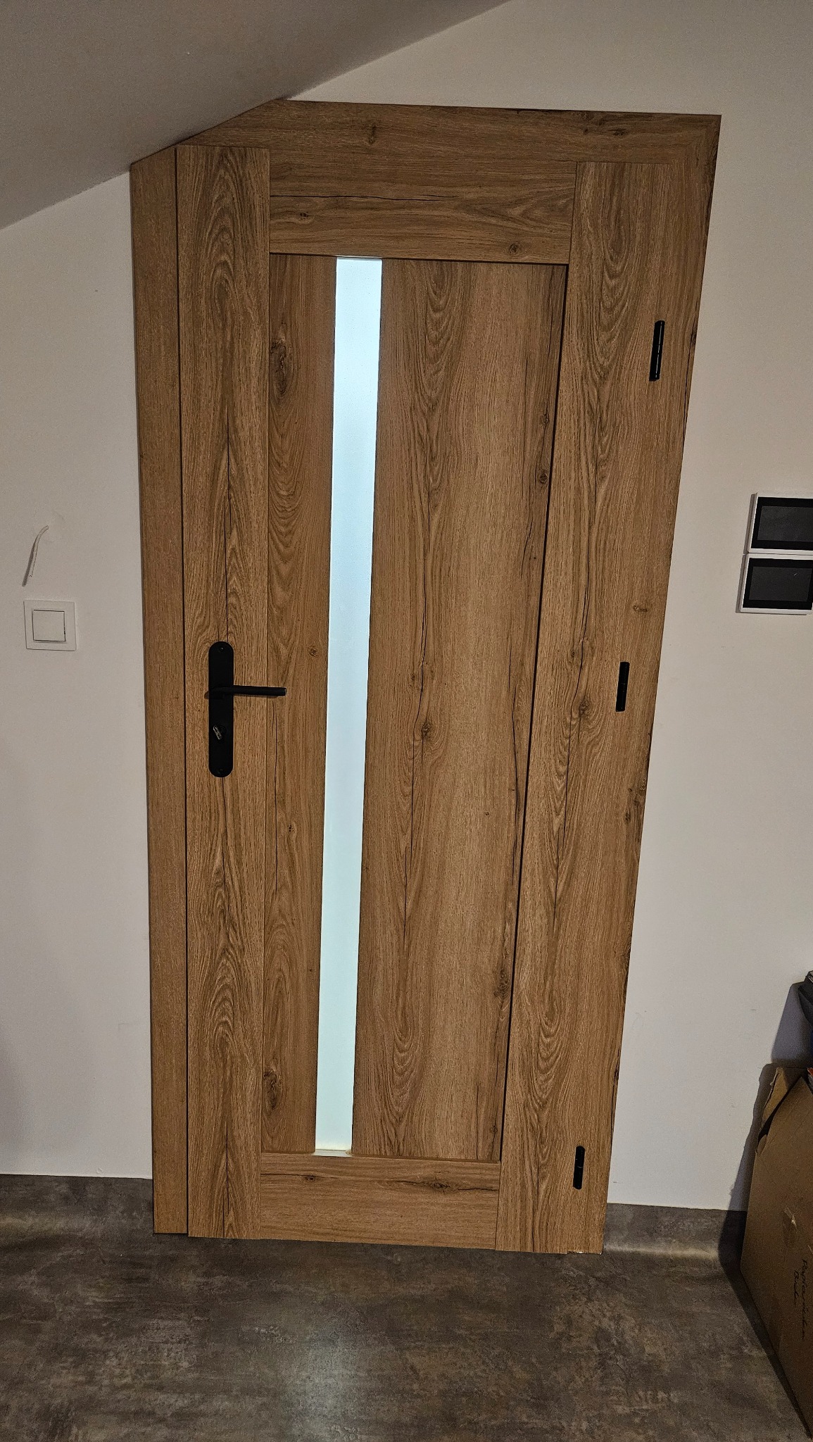 Zamknięte, brązowe drzwi wewnętrzne z pionowym, matowym przeszkleniem, czarną klamką i ukrytymi zawiasami, zamontowane w białej ścianie.