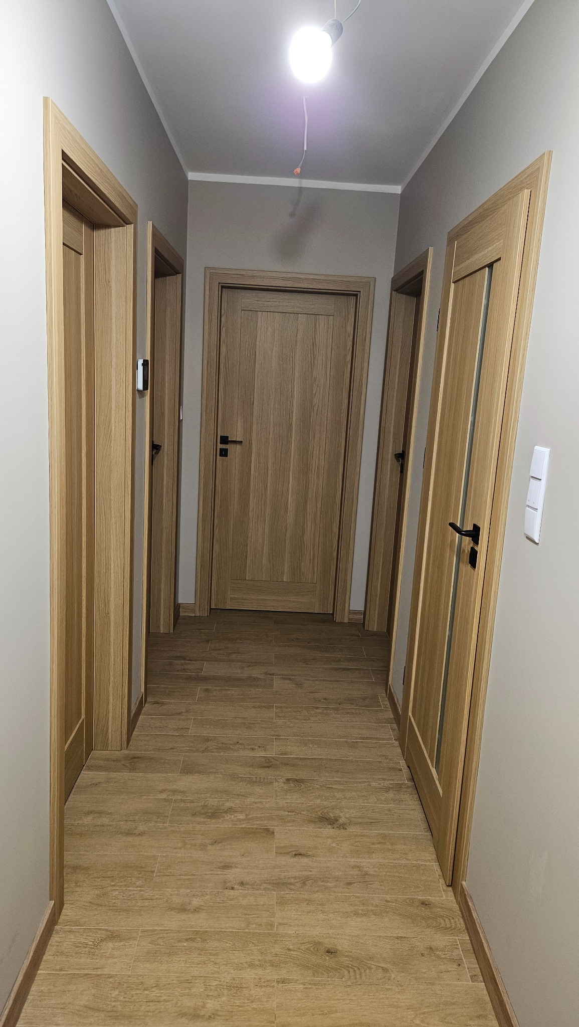 Korytarz z trzema zamontowanymi drzwiami z jasnego drewna i czarnymi klamkami, podłoga wyłożona płytkami imitującymi drewno, sufit z wiszącą żarówką bez klosza.