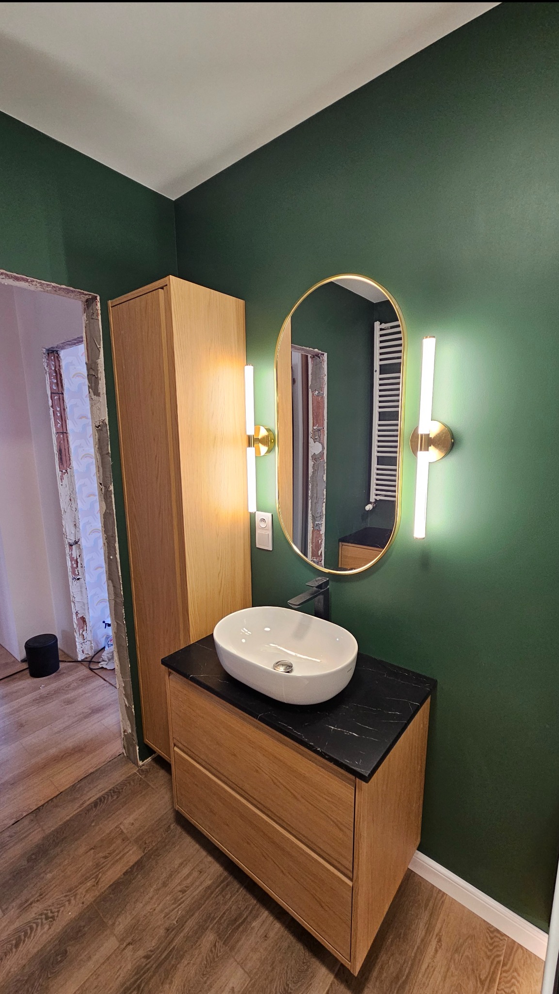 Nowoczesna łazienka z umywalką nablatową na drewnianej szafce z czarnym blatem, owalne lustro z podświetleniem LED na zielonej ścianie, widoczne prace wykończeniowe przy ościeżnicy drzwi.