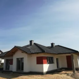 Nowoczesny dom jednorodzinny z ciemną dachówką ceramiczną i dwoma kominami, jeszcze w trakcie wykończenia terenu wokół budynku.