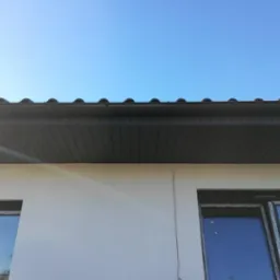 Nowoczesne, ciemnoszare podbitki dachowe z widocznym fragmentem dachu krytego dachówką ceramiczną, elewacja budynku w jasnym kolorze, błękitne niebo.