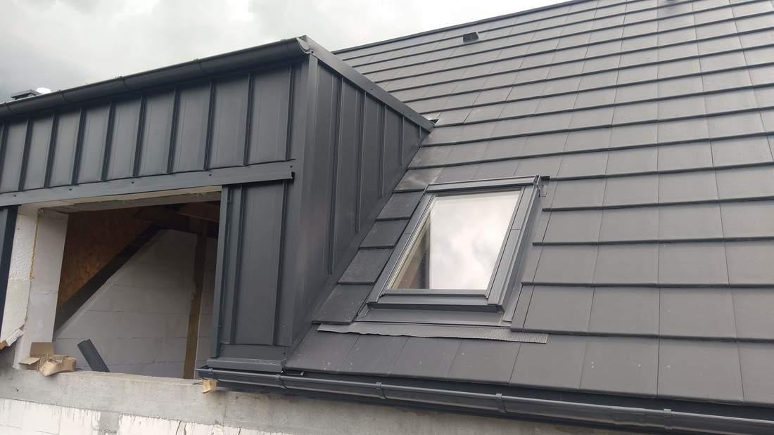 Szary dach z dachówki ceramicznej i blachy trapezowej, okno dachowe, widoczna konstrukcja poddasza w stanie surowym.