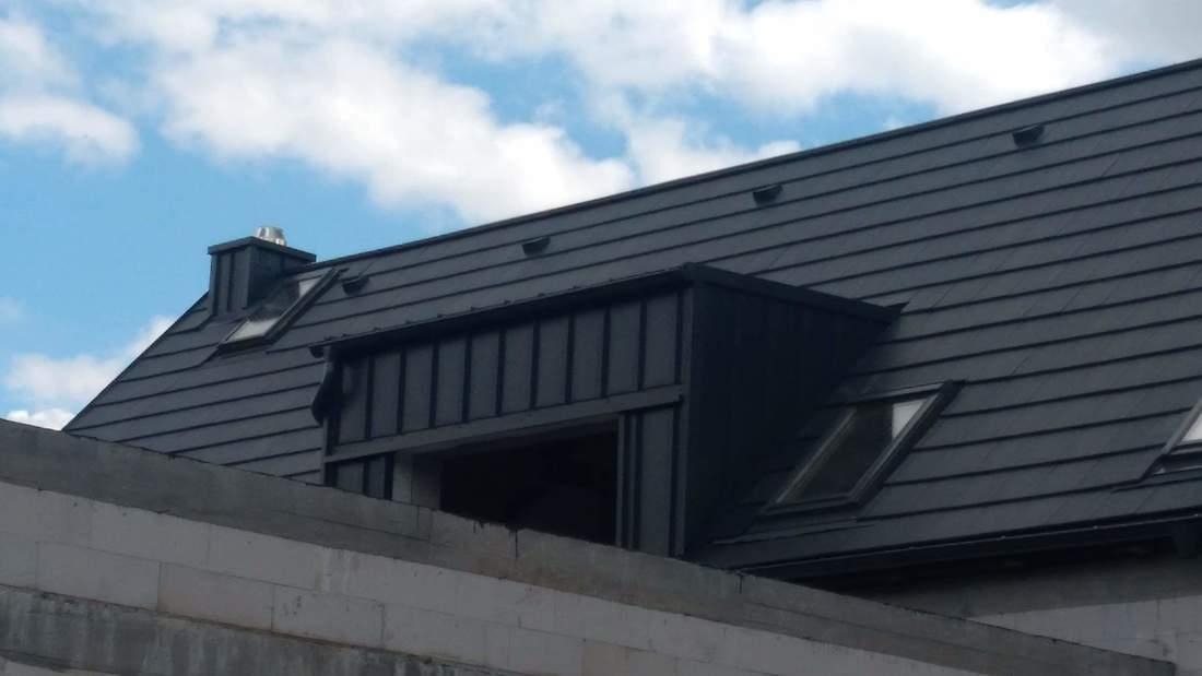 Nowy, grafitowy dach z blachy na domu w trakcie budowy, widoczne okna dachowe i komin na tle błękitnego nieba z chmurami.