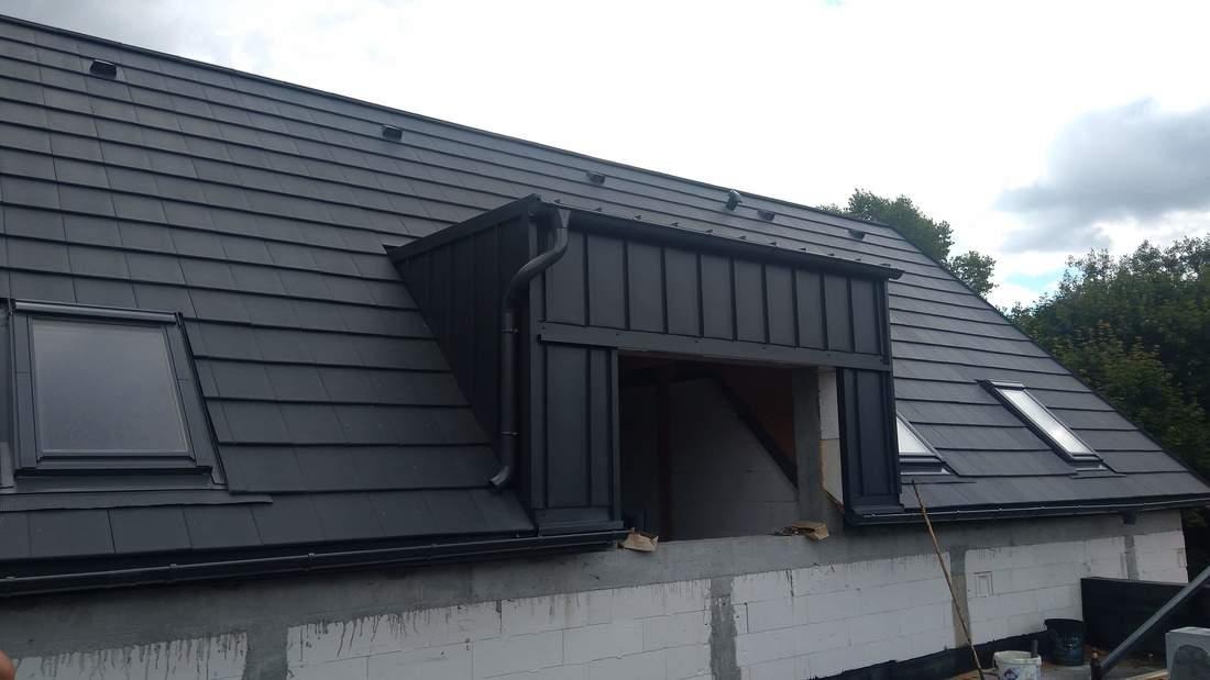 Nowy, grafitowy dach z dachówki ceramicznej z oknami połaciowymi i obróbką blacharską lukarny, widoczny fragment ściany szczytowej budynku w trakcie budowy.