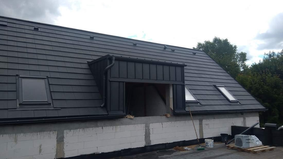 Dach pokryty grafitową dachówką ceramiczną z dwoma oknami dachowymi i lukarną w trakcie montażu, widoczne bloczki betonowe i materiały budowlane na dachu, pochmurne niebo w tle.
