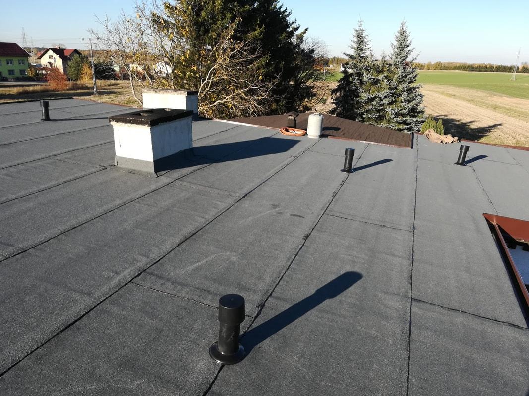 Widok z góry na płaski dach pokryty papą termozgrzewalną, z kominami, odpowietrznikami i widocznymi łączeniami, w tle zabudowania mieszkalne i pole.