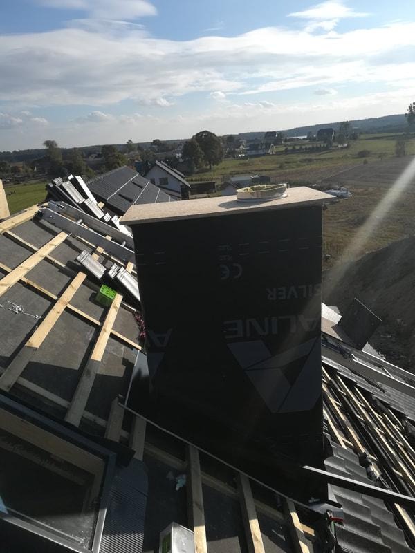 Konstrukcja dachu w trakcie budowy z widocznym kominem obłożonym czarną membraną, drewnianą konstrukcją łat i kontrłat oraz ułożonymi dachówkami w tle krajobraz wiejski.