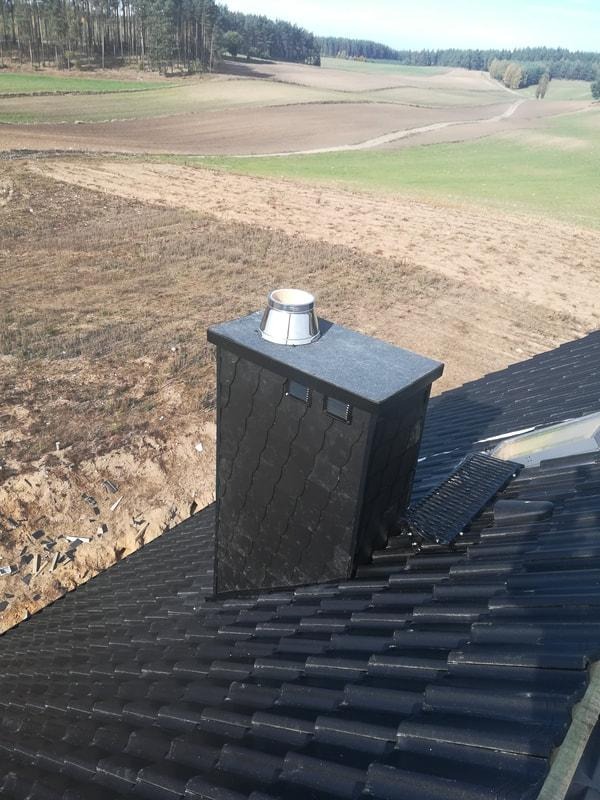 Czarny, nowy dach z dachówki ceramicznej i komin obłożony łupkiem na tle pól i lasu.
