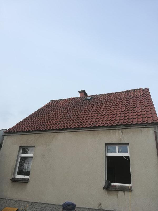 Dom w Chojnicach z dachem pokrytym czerwoną dachówką, widoczne dwa okna, jedno z nich bez szyby. Na dachu widoczny komin.