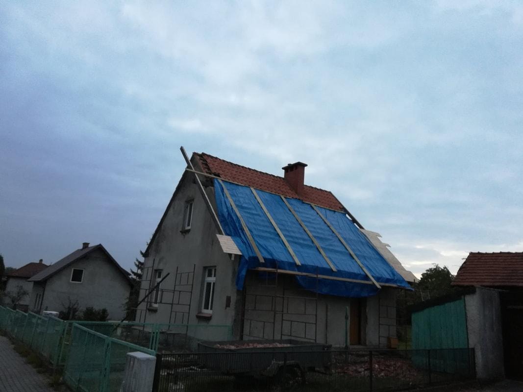 Dom jednorodzinny w trakcie prac dekarskich, zabezpieczony niebieską plandeką, z widocznymi elementami rusztowania i fragmentem starego pokrycia dachowego.