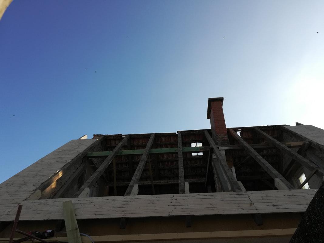 Konstrukcja dachu w trakcie demontażu, widoczne krokwie, łaty i komin na tle błękitnego nieba.