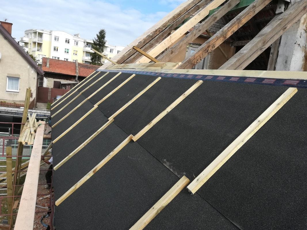 Widok z góry na konstrukcję dachu z zamontowaną membraną dachową i łatami, w tle budynki mieszkalne i błękitne niebo.