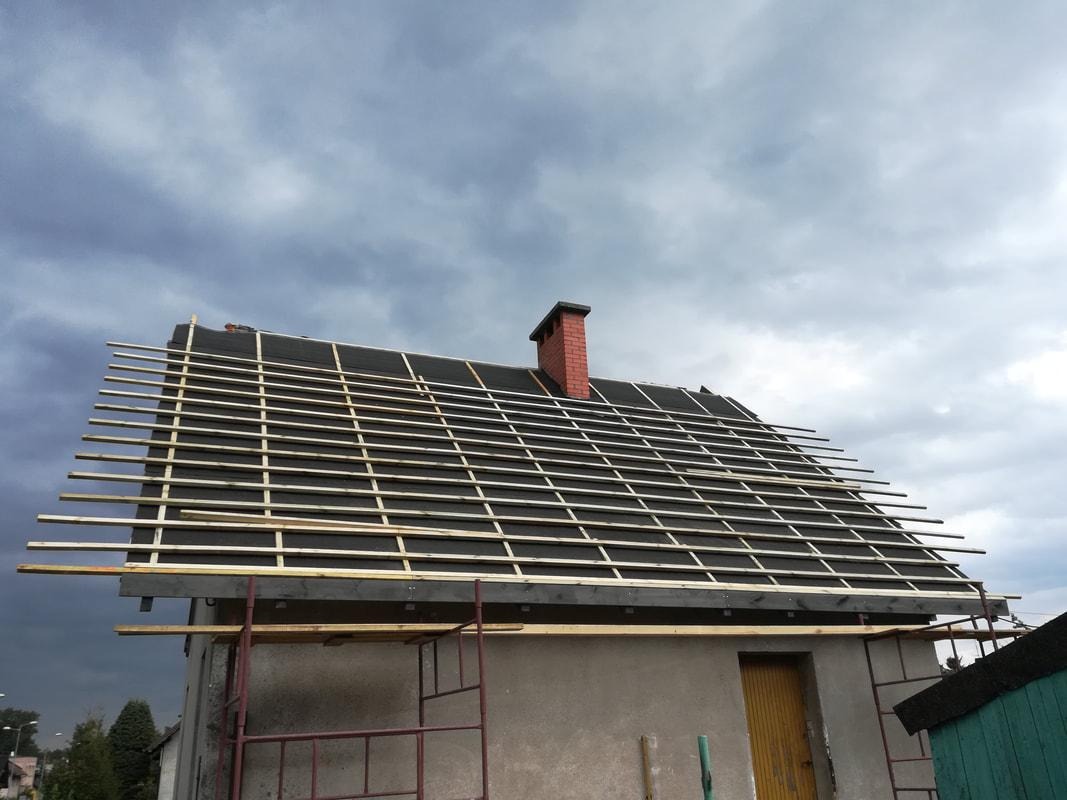 Dom jednorodzinny w trakcie wymiany dachu: widoczne deski łat na czarnej membranie dachowej, ceglany komin i fragment rusztowania na tle pochmurnego nieba.