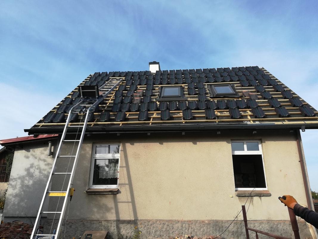 Częściowo pokryty dach z zamontowanymi oknami dachowymi, widoczna drabina dekarska i przygotowane dachówki na łatach.