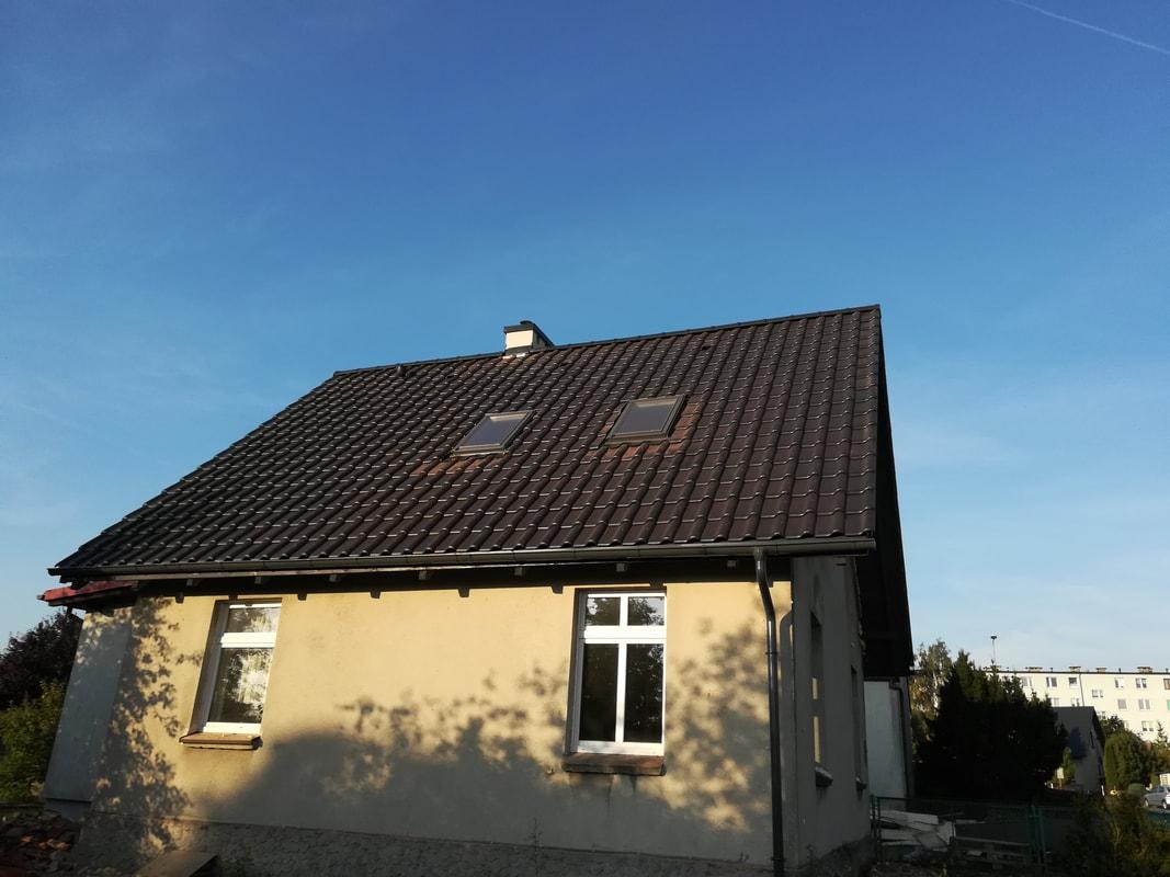 Dom jednorodzinny z nowym, brązowym dachem z dachówki ceramicznej, dwoma oknami dachowymi i rynną. Na ścianie budynku widoczne cienie drzew.