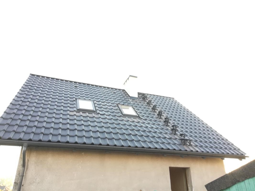 Nowy, grafitowy dach z dachówki ceramicznej z zamontowanymi oknami dachowymi i systemem przeciwśniegowym, widok z dołu na surową elewację budynku.