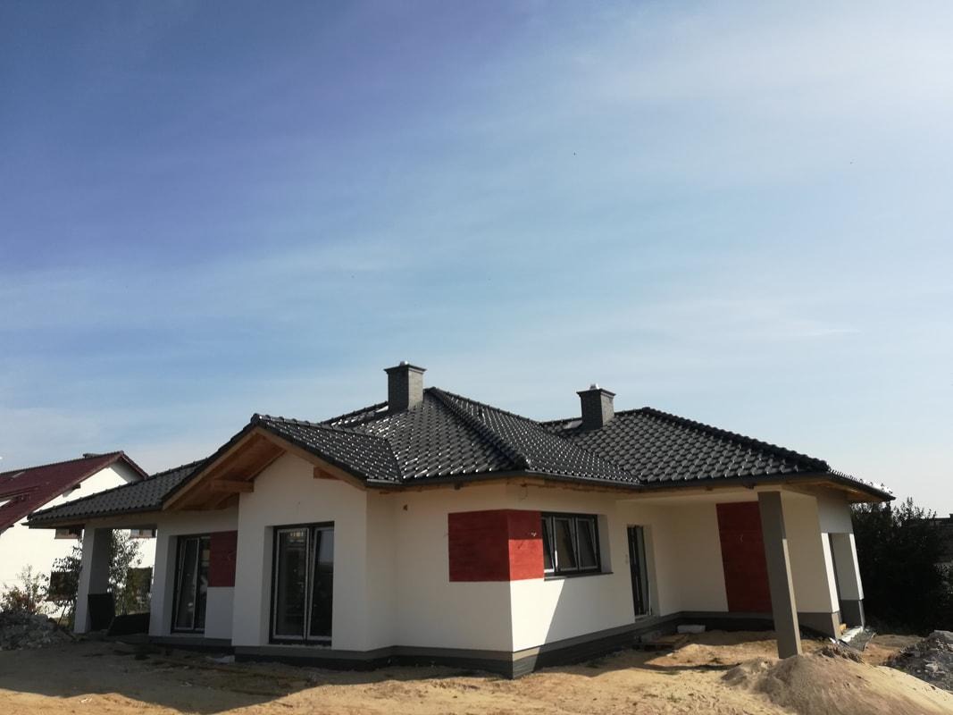 Nowoczesny dom jednorodzinny z ciemną dachówką ceramiczną i dwoma kominami, jeszcze w trakcie wykończenia terenu wokół budynku.