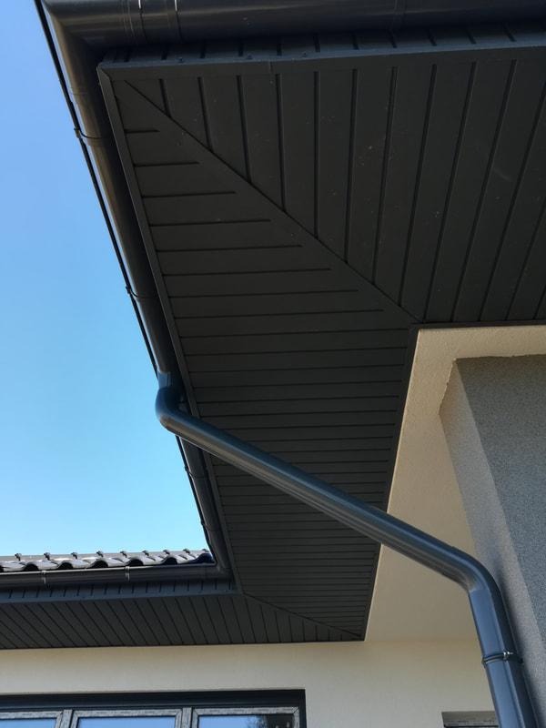 Szara rynna spustowa zamontowana na budynku z ciemnoszarą podbitką dachową i fragmentem dachu z dachówką bitumiczną, błękitne niebo w tle.