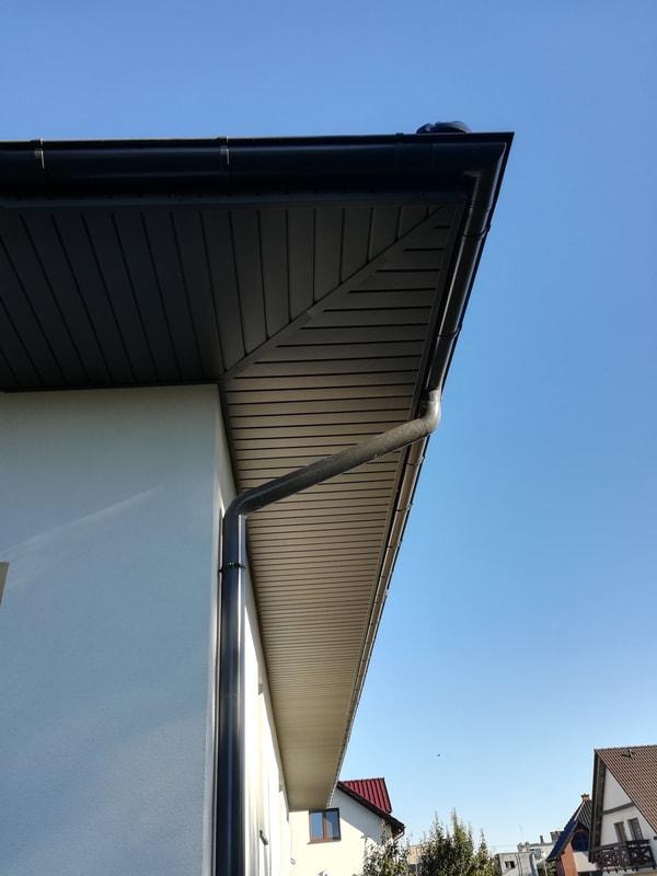 Czarne rynny spustowe i obróbki blacharskie na narożniku dachu domu jednorodzinnego z widocznym podsufitką i błękitnym niebem w tle.