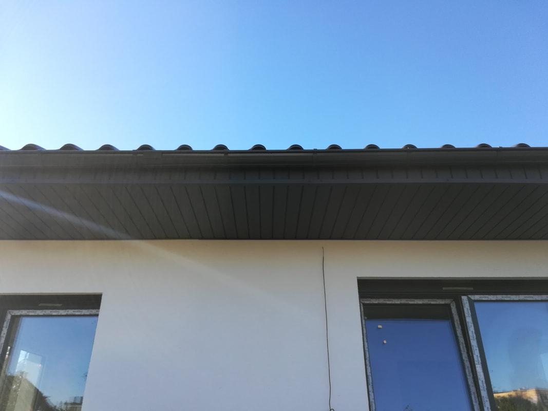 Nowoczesne, ciemnoszare podbitki dachowe z widocznym fragmentem dachu krytego dachówką ceramiczną, elewacja budynku w jasnym kolorze, błękitne niebo.