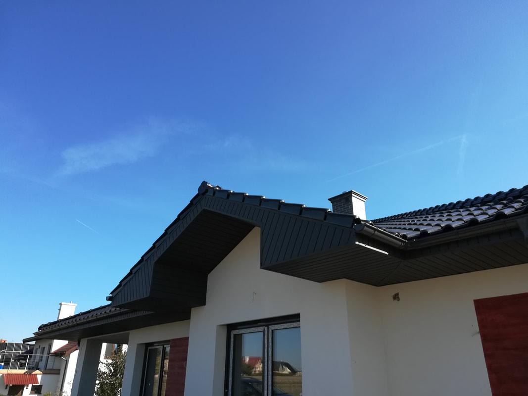 Nowoczesny dom jednorodzinny z ciemnoszarą dachówką ceramiczną i rynnami w kolorze dachu, na tle błękitnego nieba.