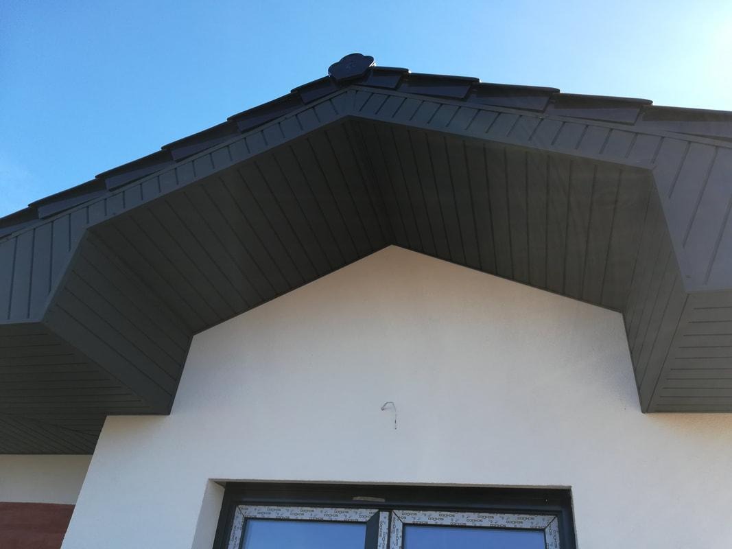 Fragment domu z nowym, ciemnoszarym dachem z dachówki ceramicznej i podbitką, widok z dołu na tle błękitnego nieba.