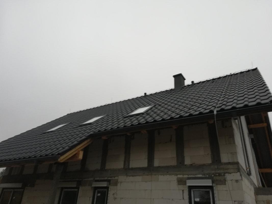 Nowo położony, ciemnoszary dach z dachówki ceramicznej z dwoma oknami dachowymi na budynku w trakcie budowy, widoczne surowe ściany z pustaków i fragmenty drewnianej konstrukcji. Zamontowane rynny...